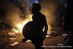 Person with helmet and wooden shield beside fire at protest 4AoaW0