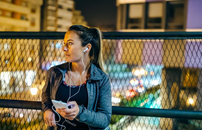 Female athlete with earphones looking away while resting over bridge after training at night in town
