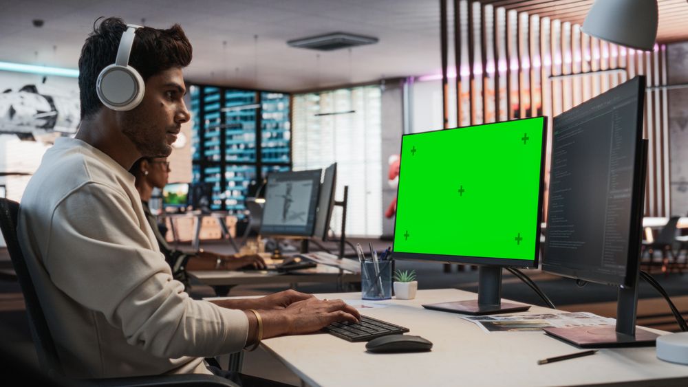 Portrait of Creative Indian Man Sitting at a Desk Using Desktop Computer with Mock-up Green Screen Chromakey.