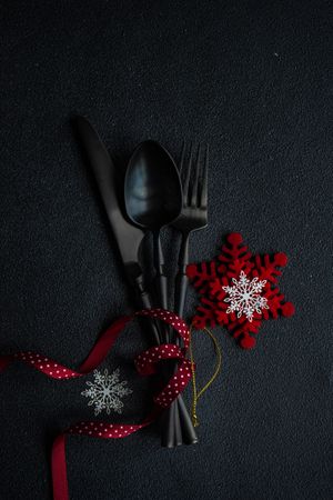 Dark knife and fork on dark background tied with festive red ribbon and snowflake ornament