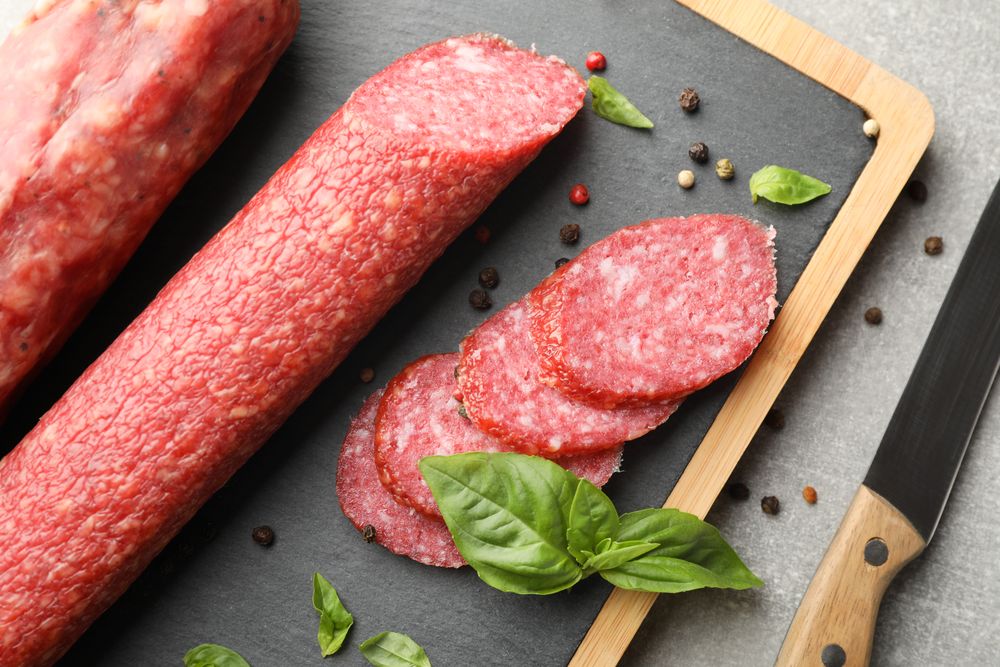 Looking down at two sticks of cured meats with slices on wooden board, closeup