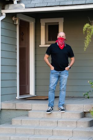 Full length shot of man in casual attire and mask standing on front porch of house