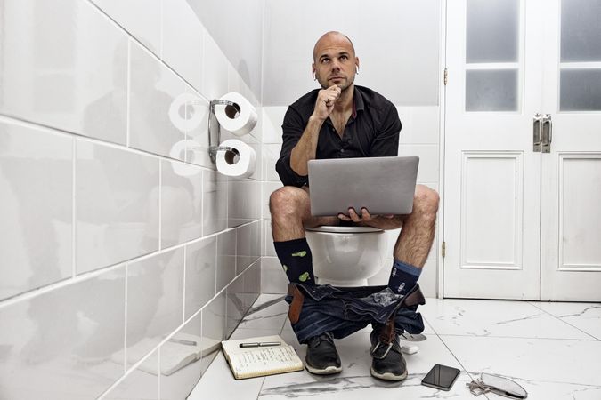 Man sitting on a toilet seat holding a laptop