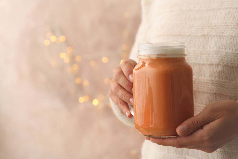 Woman hold glass jar of pumpkin latte against brown background with blurred lights