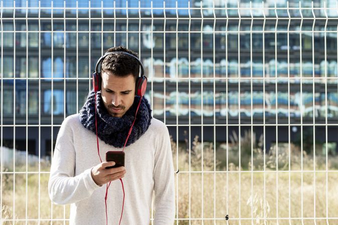 Young man walking in scarf leaning back on metallic fence and looking down at smartphone