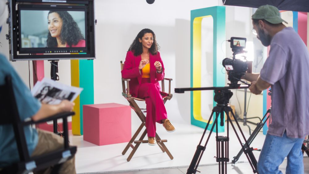 Young Woman In A Vibrant Red Suit Speaks Animatedly On A Director's Chair, Surrounded By Colorful Set Design, With A Male Cinematographer Recording For A Lively Television Or Web Broadcast.
