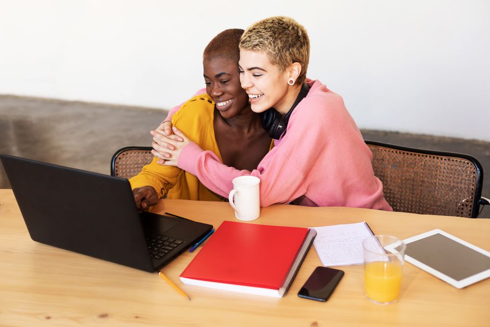 Female friends embracing happily over laptop at breakfast table
