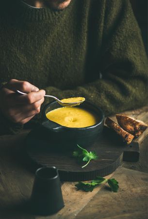 Man with stubble eating warm yellow soup from dark bowl with toast