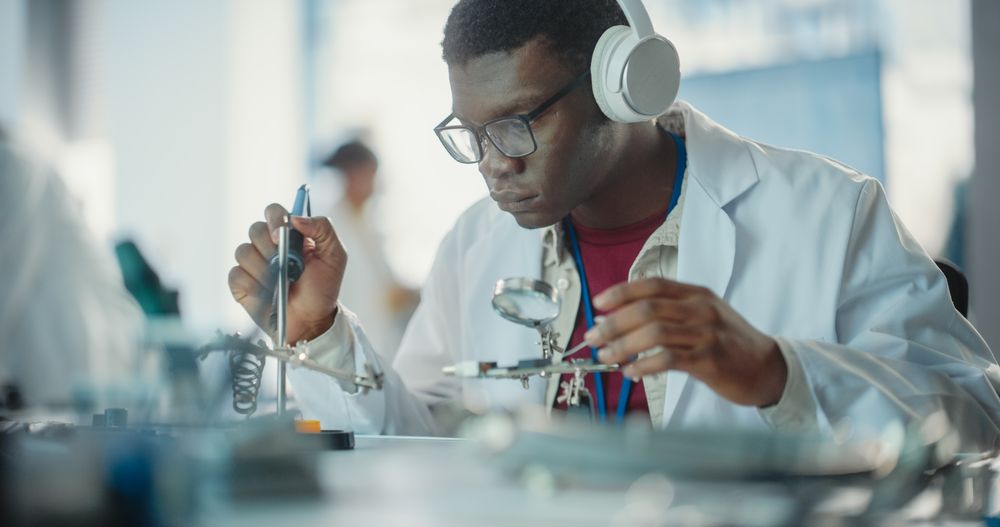 Portrait of Black Male Lab Specialist Soldering a Circuit Board, Using Headphones in a Modern Industrial Company.