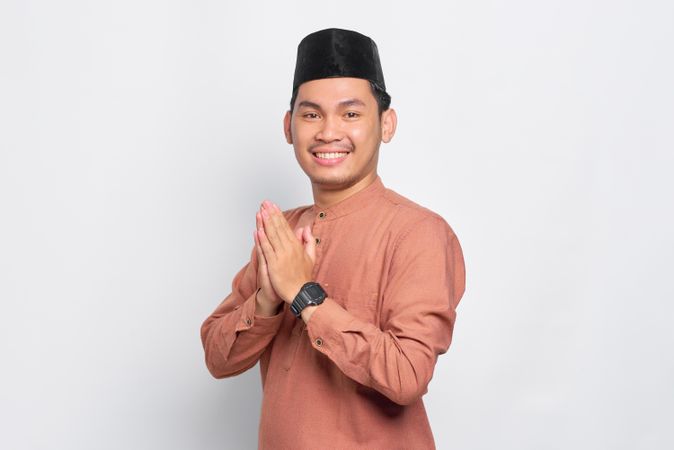 Muslim man in kufi hat smiling with hands closed in prayer standing to the side