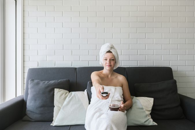 Happy relaxed woman enjoying day off while having hot drink after shower and watching TV in apartment in loft style