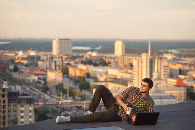 Man reclining on roof over looking city working on laptop with takeaway drink