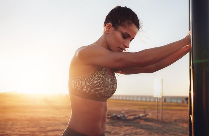 Shot of muscular young woman wearing sports bra standing on the beach with sun setting behind