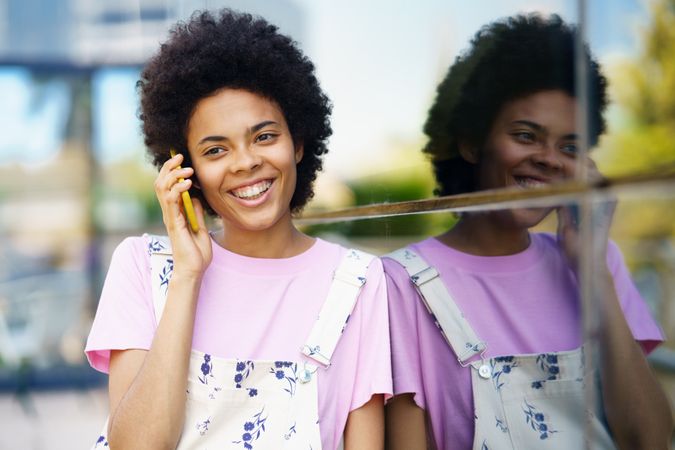 Happy female in floral overalls speaking on cell phone and leaning on window outside