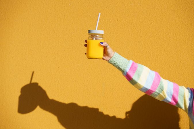 Person in striped sleeve holding cup of orange juice on yellow wall