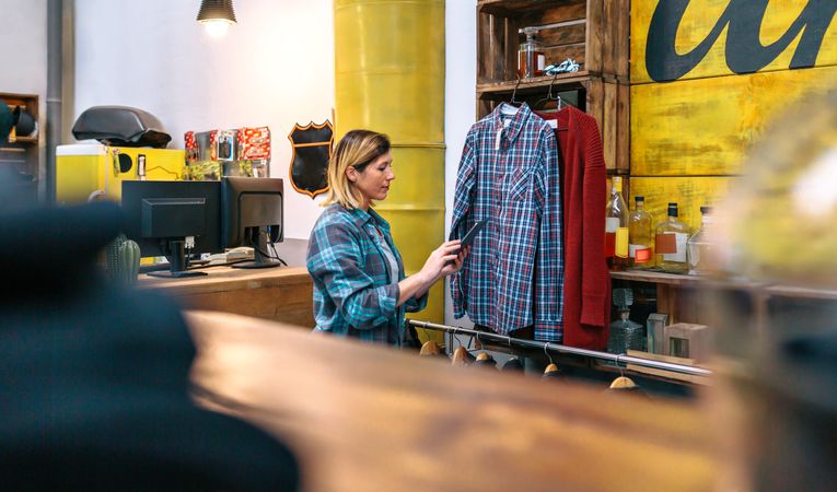 Blonde young woman looking digital tablet while checking stock on second hand clothing store