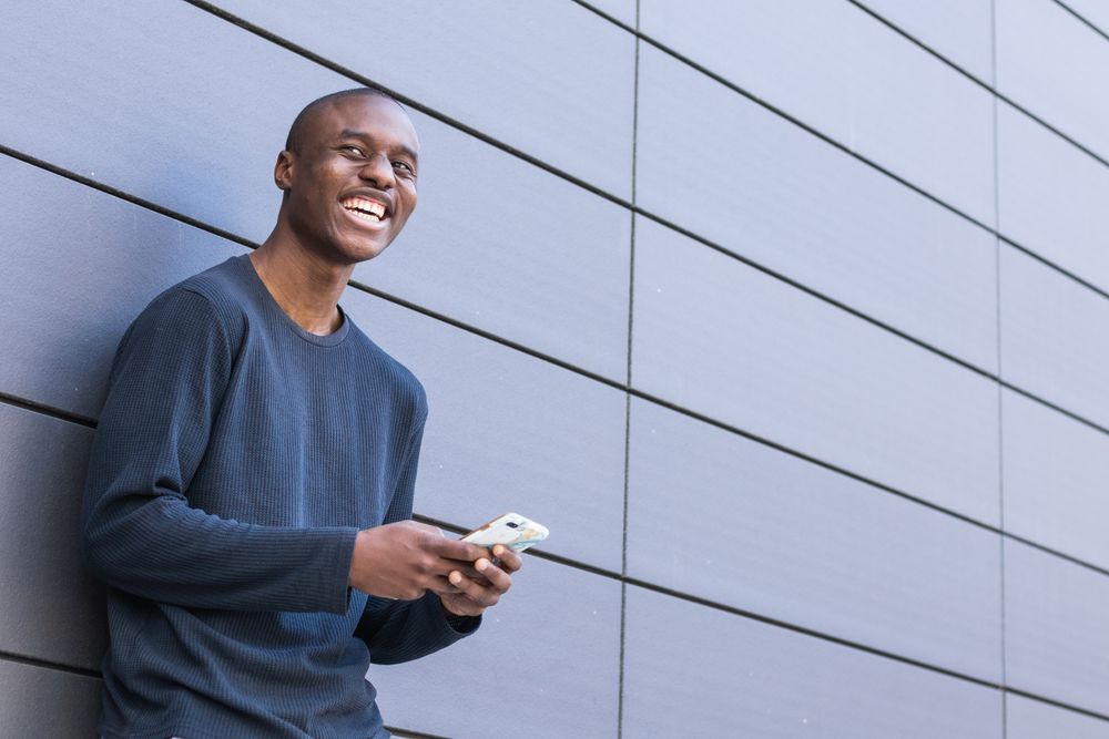 Happy Black man using smartphone against gray wall