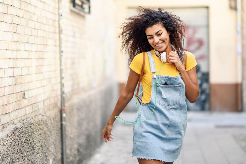 Dancing Arab woman with headphones and curly hairstyle walking outdoors in lane with graffiti