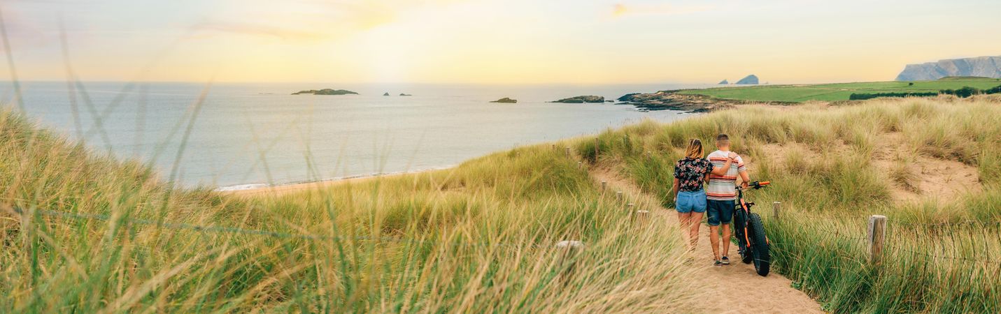 Back of couple strolling with bicycle on sandy path towards the ocean