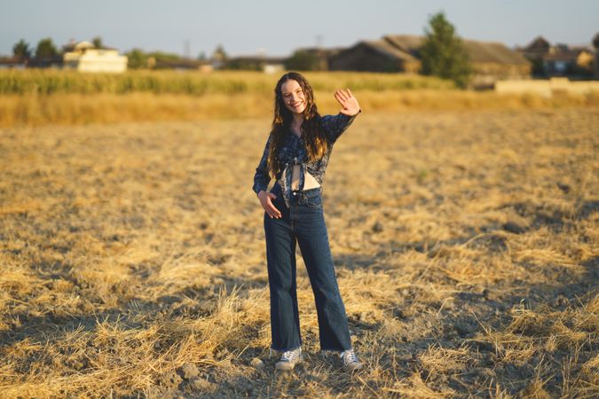 A Young Woman Dressed in a Stylish Denim Outfit Waves Cheerfully in a Beautiful Golden Field
