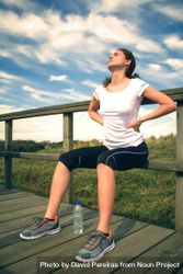 Woman in athletic gear holding hurt back while seated on wooden path 5rdm7b