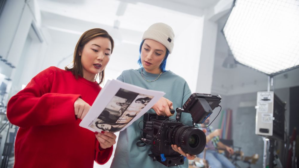 In A Modern Studio, An Asian Woman In A Red Sweater And A Woman With Blue Hair, Both Young, Collaborate Over A Camera And Storyboard, Focusing On Creative Film Production And Advertising.