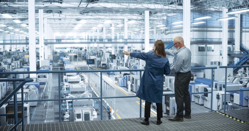 Experienced Male and Female Engineers Standing on a Platform with Their Back to Camera, Using Laptop Computer and Discussing Production at a Modern AI Automated Electronics Manufacture