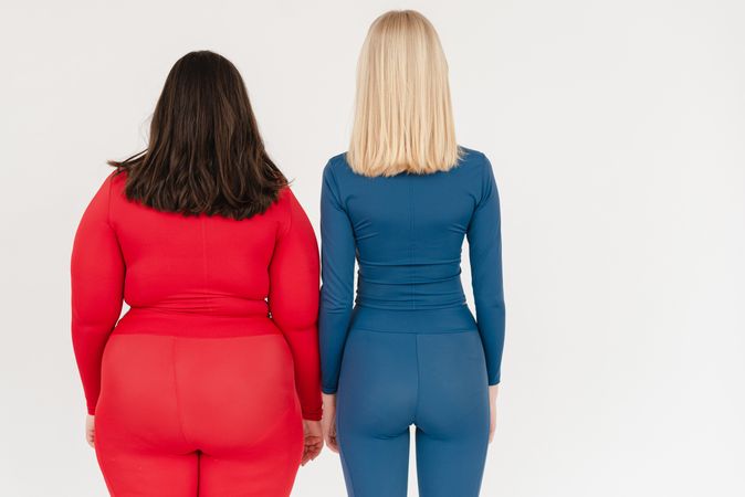 Back view woman in blue outfit standing beside woman in red outfit against light background