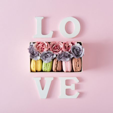 The word “Love” surrounding box of macarons and flowers on pink background