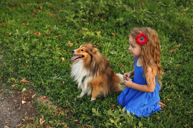 Girl in blue dress with flower in hair sitting with dog in the grass