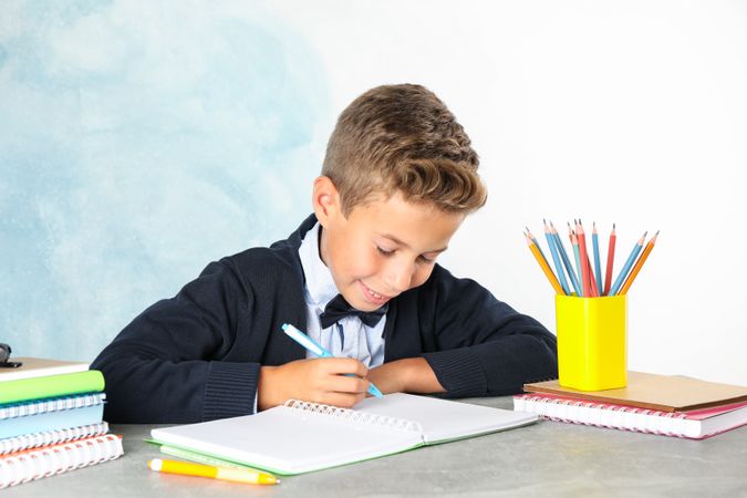 Boy in school uniform writes sitting at the table against color background