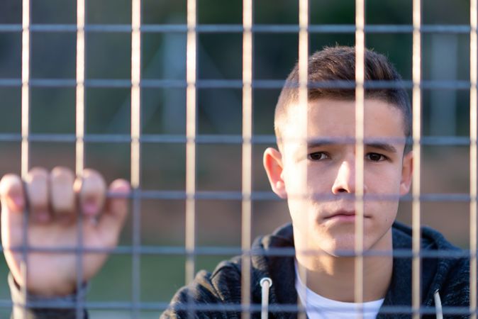 Teenage male in hoodie standing behind fence looking at camera