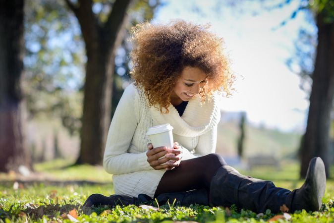 Woman laughing and looking down at warm drink sitting in park