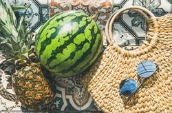 Thatched bag on pattered tiles with sunglasses, watermelon and pineapple, horizontal composition
