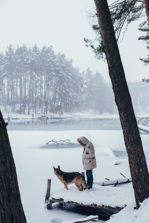 Man in parka outside looking out onto lake in snowy forest with dog at his side