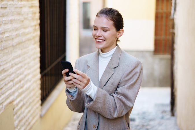 A cheerful young woman in a stylish outfit smiles while using her smartphone in a city alley