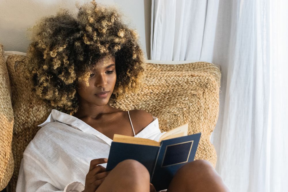 Young Black woman sitting on bed and reading book