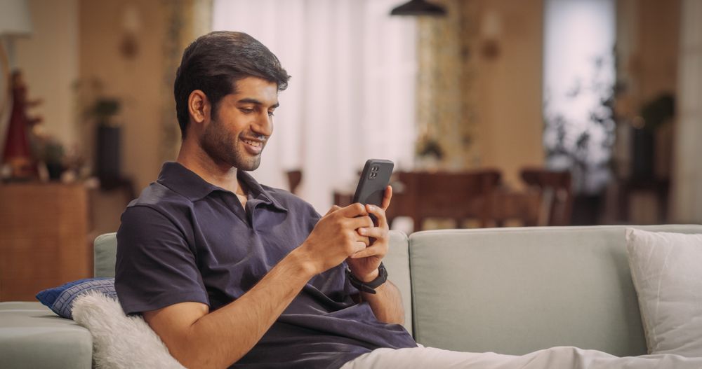 Indian Man Lying on Sofa Using Smartphone, Exploring Online Shopping