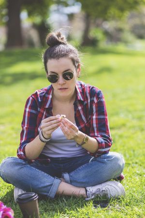 Front view of a young hipster woman sitting on grass in a park while holding a flower in a sunny day
