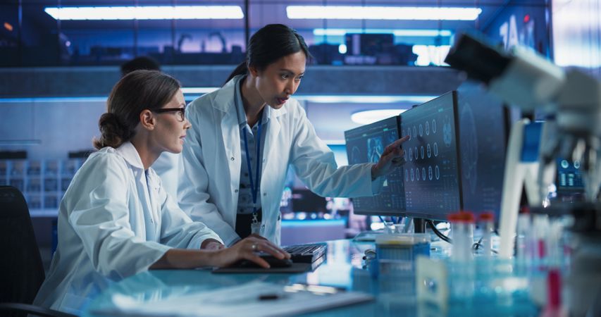Asian And White Female Doctors Talking In Medical Research Center And Using Desktop Computer To Analyze MRI Scans Of Brain.