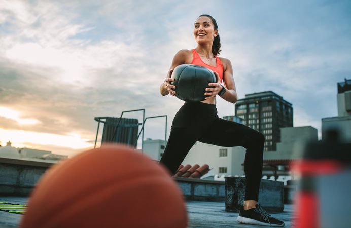 Female athlete doing forward lunges holding a medicine ball on rooftop