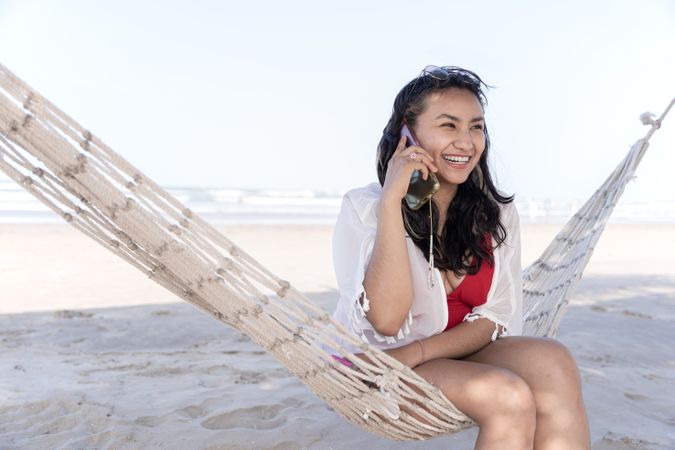 A young Hispanic woman is smiling while speaking on her phone on a hammock at the beach