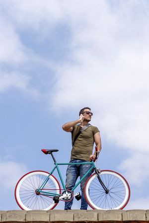 Male speaking on phone and standing with bike on roof with beautiful blue sky and clouds, vertical