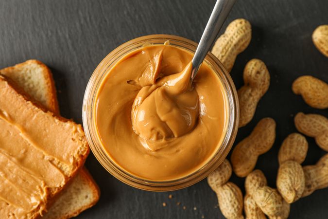 Glass jar with peanut butter and spoon, peanut, peanut butter sandwich and cutting board on wooden background, space for text and closeup