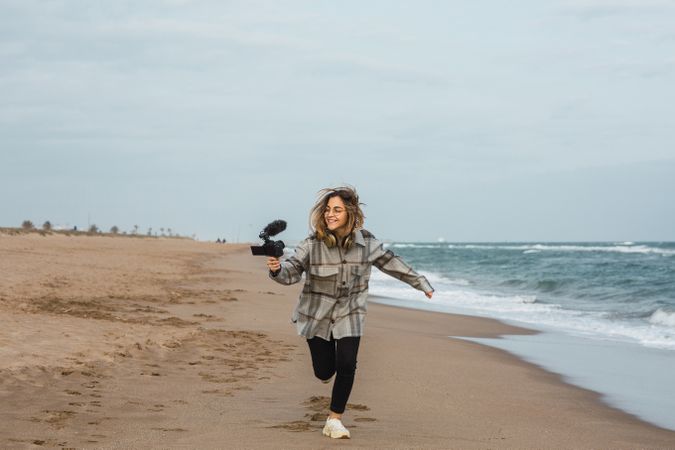 Cheerful young woman running on seashore and shooting video on camera