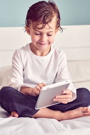 Smiling boy with wet hair browsing tablet on bed