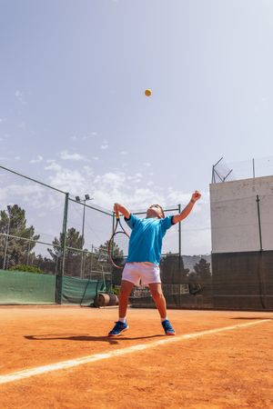 Little boy playing tennis on a dirt court making a serve