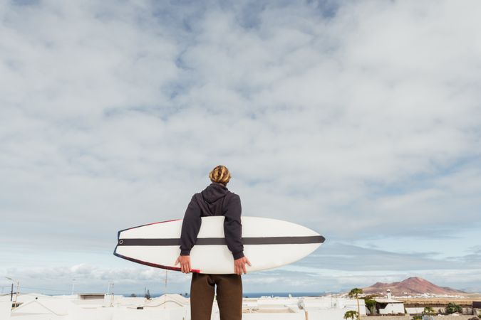 Man standing with surf board on roof of building and looking at mountain