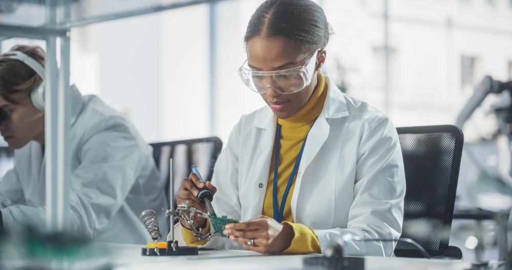 Black Female Working with Soldering Iron Wearing Safety Goggles and Lab Coat