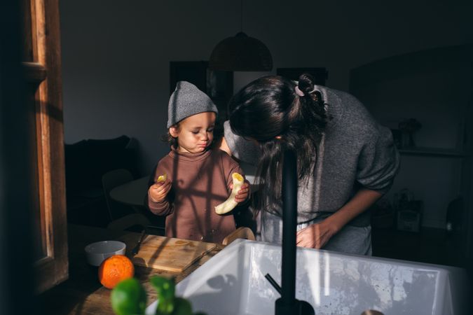 Young mother embracing little son eating banana in kitchen